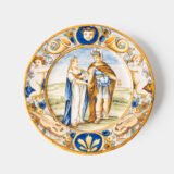 Hand-painted Italian maiolica plate, Deruta or Urbino, late 17th to early 18th century. Depicts Mercury with winged sandals beside a female figure, likely an allegory of Peace, Concord, or Wisdom. Rich Baroque border with cherubs, musical instruments, and cartouches. Tin-glazed earthenware with visible craquelure and minor rim losses.