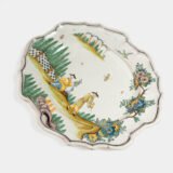 Oval faience plate, France, ca. 1760–1790, attributed to Rouen; tin-glazed earthenware with hand-painted chinoiserie scene in enamel pigments (cobalt, manganese, copper, antimony); figures in stylized garden within manganese-lined scalloped rim; ivory glaze with fine, stable craquelure