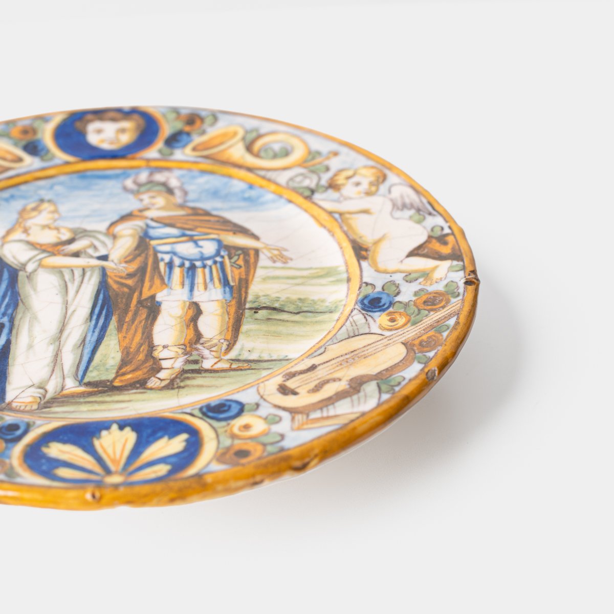 Hand-painted Italian maiolica plate, Deruta or Urbino, late 17th to early 18th century. Depicts Mercury with winged sandals beside a female figure, likely an allegory of Peace, Concord, or Wisdom. Rich Baroque border with cherubs, musical instruments, and cartouches. Tin-glazed earthenware with visible craquelure and minor rim losses.
