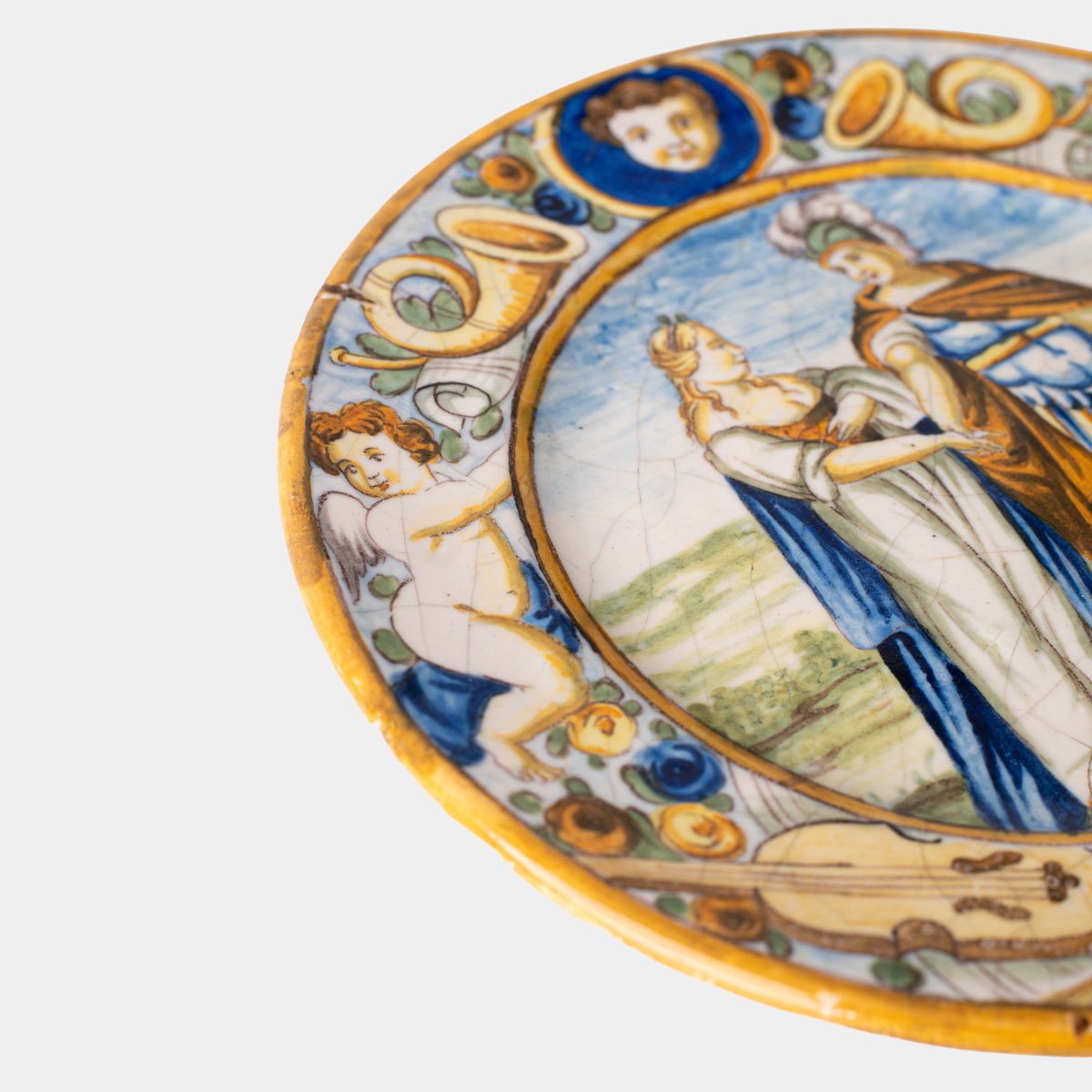 Hand-painted Italian maiolica plate, Deruta or Urbino, late 17th to early 18th century. Depicts Mercury with winged sandals beside a female figure, likely an allegory of Peace, Concord, or Wisdom. Rich Baroque border with cherubs, musical instruments, and cartouches. Tin-glazed earthenware with visible craquelure and minor rim losses.