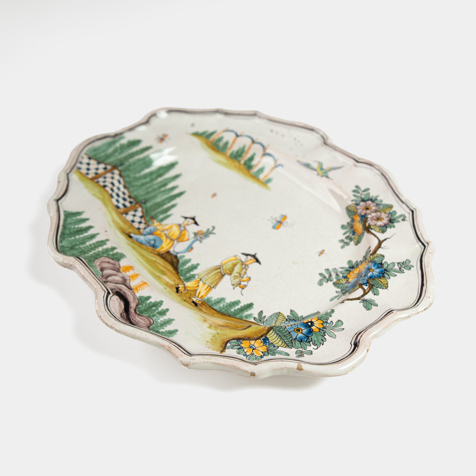 Oval faience plate, France, ca. 1760–1790, attributed to Rouen; tin-glazed earthenware with hand-painted chinoiserie scene in enamel pigments (cobalt, manganese, copper, antimony); figures in stylized garden within manganese-lined scalloped rim; ivory glaze with fine, stable craquelure