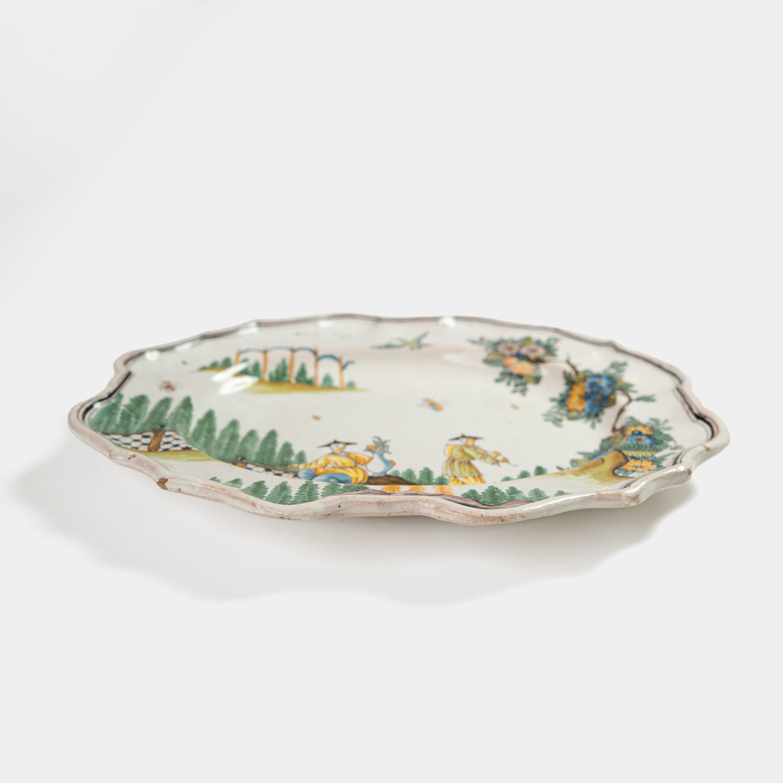 Oval faience plate, France, ca. 1760–1790, attributed to Rouen; tin-glazed earthenware with hand-painted chinoiserie scene in enamel pigments (cobalt, manganese, copper, antimony); figures in stylized garden within manganese-lined scalloped rim; ivory glaze with fine, stable craquelure