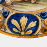 Hand-painted Italian maiolica plate, Deruta or Urbino, late 17th to early 18th century. Depicts Mercury with winged sandals beside a female figure, likely an allegory of Peace, Concord, or Wisdom. Rich Baroque border with cherubs, musical instruments, and cartouches. Tin-glazed earthenware with visible craquelure and minor rim losses.