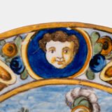 Hand-painted Italian maiolica plate, Deruta or Urbino, late 17th to early 18th century. Depicts Mercury with winged sandals beside a female figure, likely an allegory of Peace, Concord, or Wisdom. Rich Baroque border with cherubs, musical instruments, and cartouches. Tin-glazed earthenware with visible craquelure and minor rim losses.