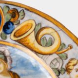 Hand-painted Italian maiolica plate, Deruta or Urbino, late 17th to early 18th century. Depicts Mercury with winged sandals beside a female figure, likely an allegory of Peace, Concord, or Wisdom. Rich Baroque border with cherubs, musical instruments, and cartouches. Tin-glazed earthenware with visible craquelure and minor rim losses.