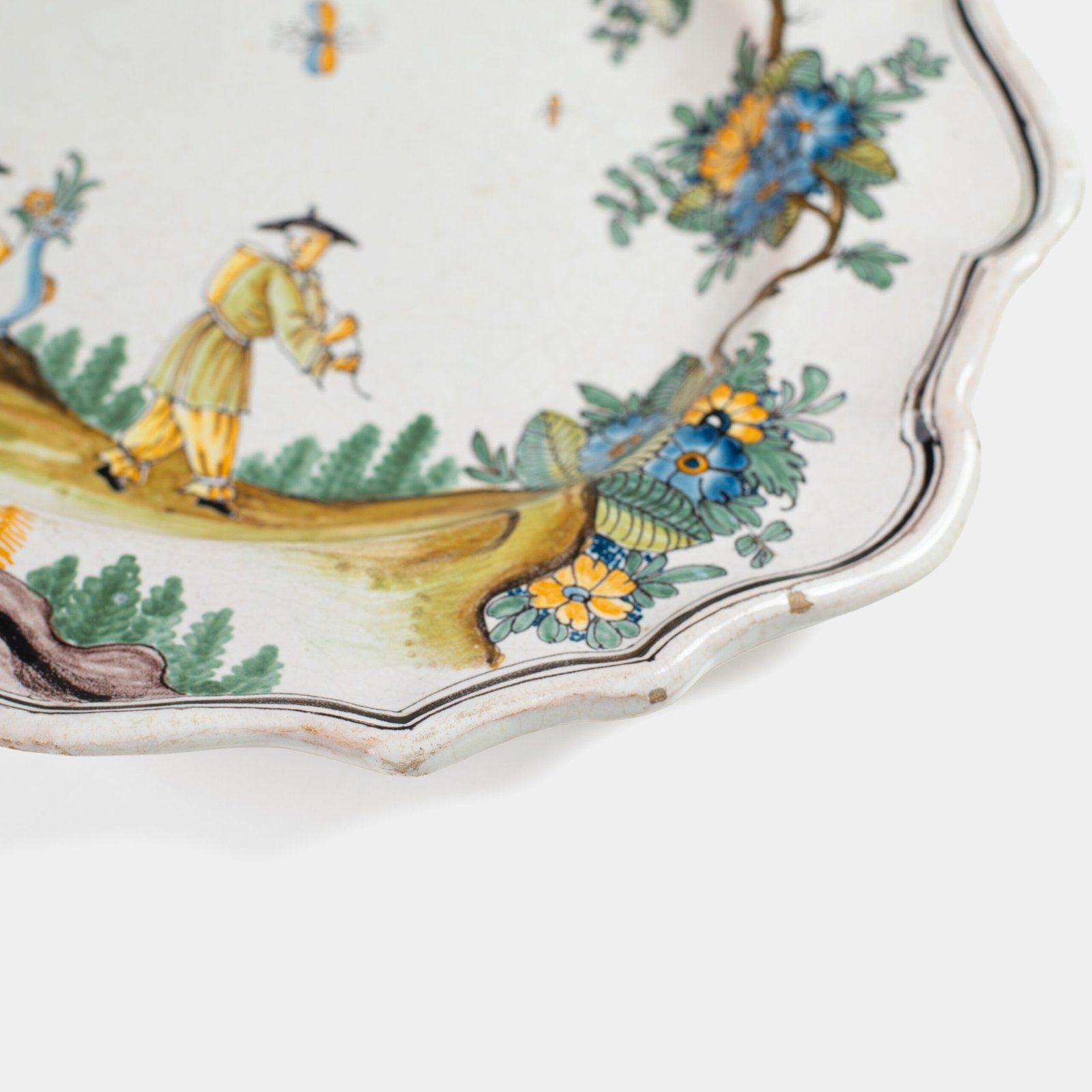 Oval faience plate, France, ca. 1760–1790, attributed to Rouen; tin-glazed earthenware with hand-painted chinoiserie scene in enamel pigments (cobalt, manganese, copper, antimony); figures in stylized garden within manganese-lined scalloped rim; ivory glaze with fine, stable craquelure