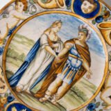 Hand-painted Italian maiolica plate, Deruta or Urbino, late 17th to early 18th century. Depicts Mercury with winged sandals beside a female figure, likely an allegory of Peace, Concord, or Wisdom. Rich Baroque border with cherubs, musical instruments, and cartouches. Tin-glazed earthenware with visible craquelure and minor rim losses.