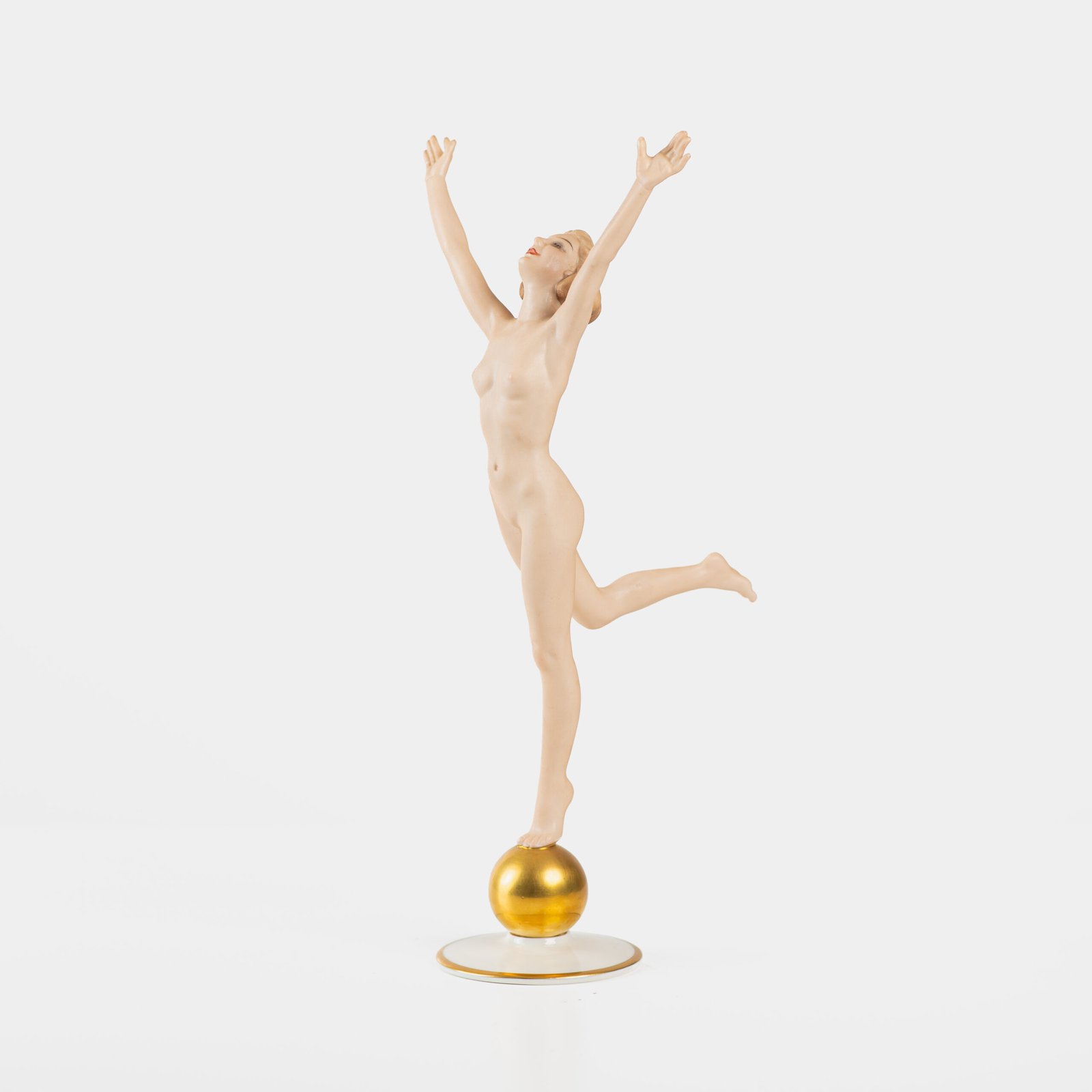 Karl Tutter porcelain figurine “Sonnenkind” – Art Deco sculpture by Hutschenreuther Germany, c.1935–1940 Art Deco porcelain figurine titled “Sonnenkind” (“Sun Child”), modeled by Karl Tutter for Hutschenreuther ca. 1935–1940. Depicts a nude young woman balancing on a golden sphere, arms extended in a dynamic pose. White porcelain with a matte pinkish flesh-tone finish, painted facial features, and satin brown hair. Gilded accents on the sphere and base. Rare signed version with circular base and gold trim.