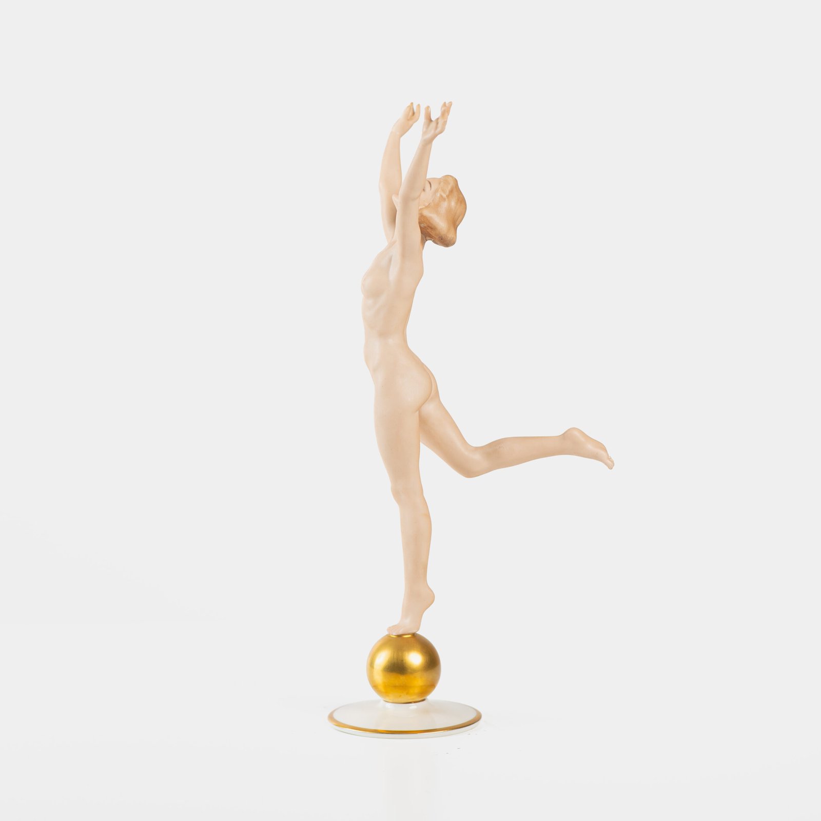 Karl Tutter porcelain figurine “Sonnenkind” – Art Deco sculpture by Hutschenreuther Germany, c.1935–1940 Art Deco porcelain figurine titled “Sonnenkind” (“Sun Child”), modeled by Karl Tutter for Hutschenreuther ca. 1935–1940. Depicts a nude young woman balancing on a golden sphere, arms extended in a dynamic pose. White porcelain with a matte pinkish flesh-tone finish, painted facial features, and satin brown hair. Gilded accents on the sphere and base. Rare signed version with circular base and gold trim.