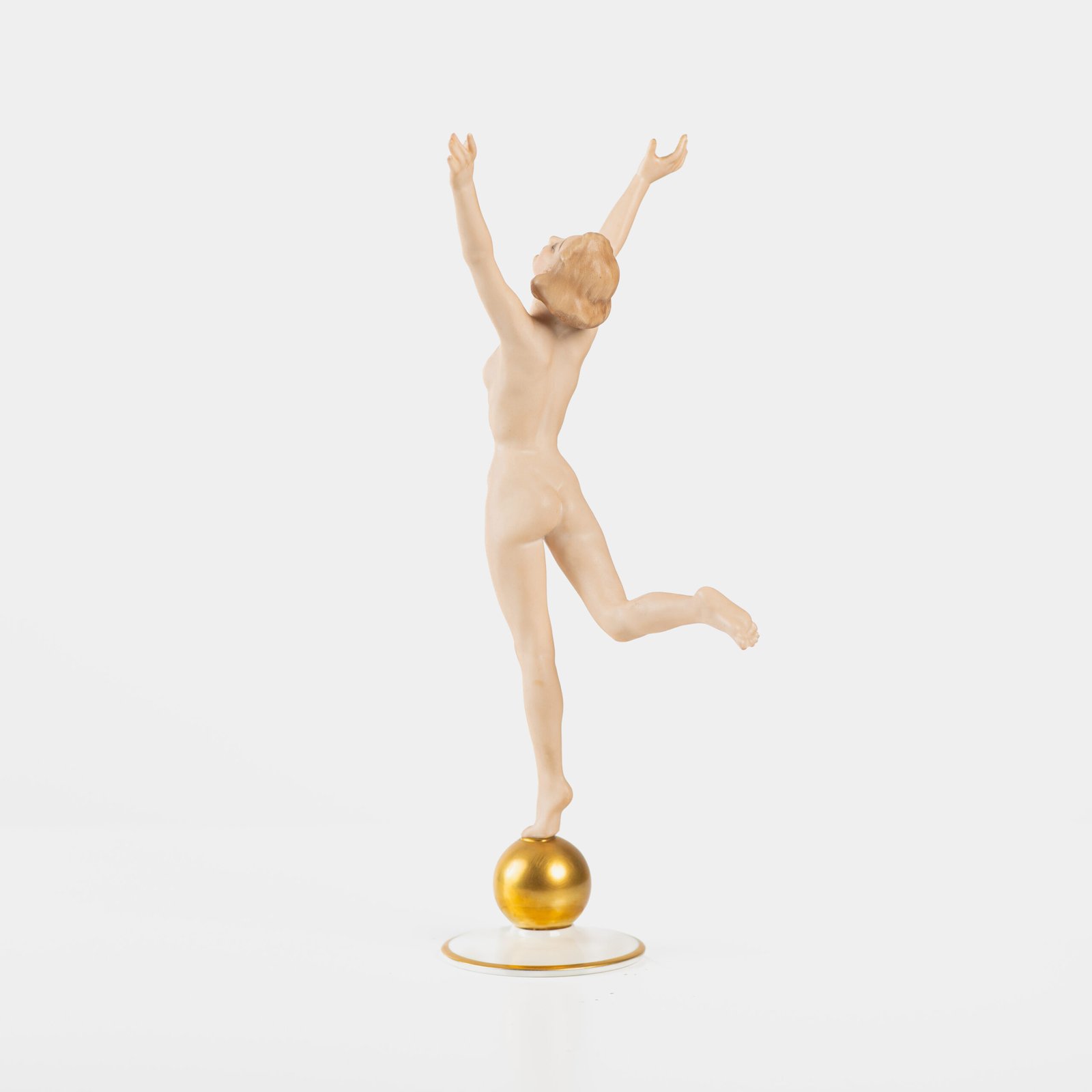 Karl Tutter porcelain figurine “Sonnenkind” – Art Deco sculpture by Hutschenreuther Germany, c.1935–1940 Art Deco porcelain figurine titled “Sonnenkind” (“Sun Child”), modeled by Karl Tutter for Hutschenreuther ca. 1935–1940. Depicts a nude young woman balancing on a golden sphere, arms extended in a dynamic pose. White porcelain with a matte pinkish flesh-tone finish, painted facial features, and satin brown hair. Gilded accents on the sphere and base. Rare signed version with circular base and gold trim.