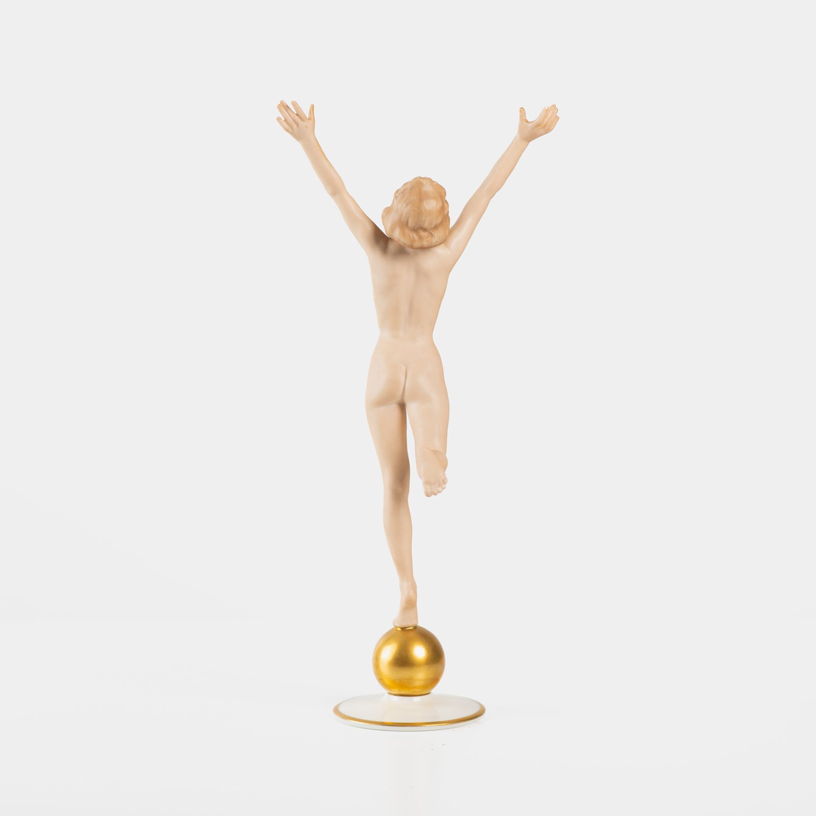 Karl Tutter porcelain figurine “Sonnenkind” – Art Deco sculpture by Hutschenreuther Germany, c.1935–1940 Art Deco porcelain figurine titled “Sonnenkind” (“Sun Child”), modeled by Karl Tutter for Hutschenreuther ca. 1935–1940. Depicts a nude young woman balancing on a golden sphere, arms extended in a dynamic pose. White porcelain with a matte pinkish flesh-tone finish, painted facial features, and satin brown hair. Gilded accents on the sphere and base. Rare signed version with circular base and gold trim.