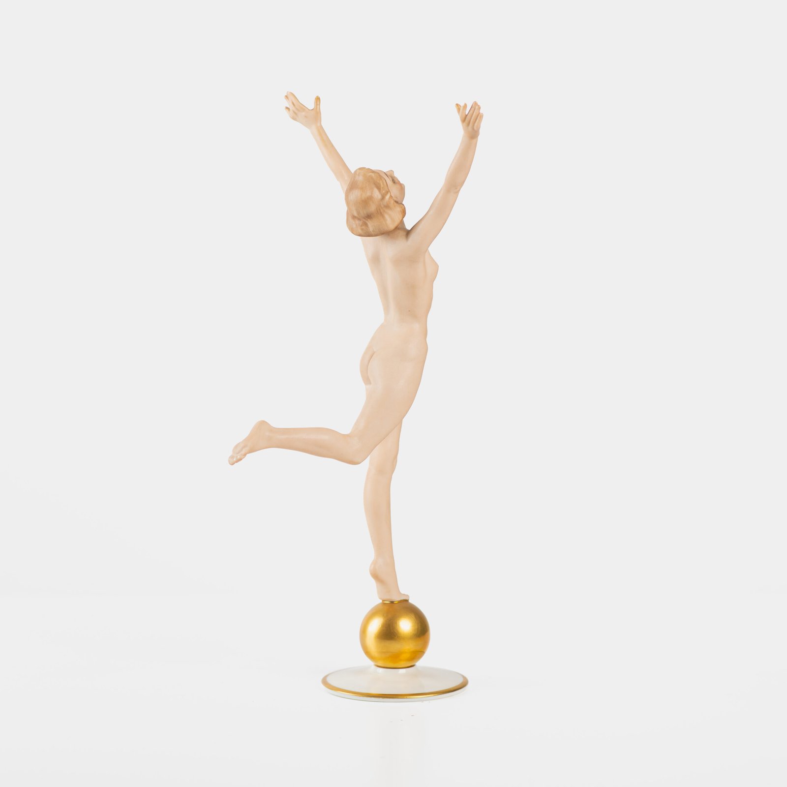 Karl Tutter porcelain figurine “Sonnenkind” – Art Deco sculpture by Hutschenreuther Germany, c.1935–1940 Art Deco porcelain figurine titled “Sonnenkind” (“Sun Child”), modeled by Karl Tutter for Hutschenreuther ca. 1935–1940. Depicts a nude young woman balancing on a golden sphere, arms extended in a dynamic pose. White porcelain with a matte pinkish flesh-tone finish, painted facial features, and satin brown hair. Gilded accents on the sphere and base. Rare signed version with circular base and gold trim.