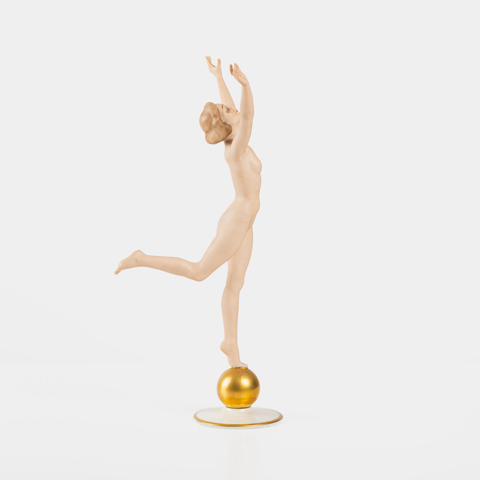 Karl Tutter porcelain figurine “Sonnenkind” – Art Deco sculpture by Hutschenreuther Germany, c.1935–1940 Art Deco porcelain figurine titled “Sonnenkind” (“Sun Child”), modeled by Karl Tutter for Hutschenreuther ca. 1935–1940. Depicts a nude young woman balancing on a golden sphere, arms extended in a dynamic pose. White porcelain with a matte pinkish flesh-tone finish, painted facial features, and satin brown hair. Gilded accents on the sphere and base. Rare signed version with circular base and gold trim.