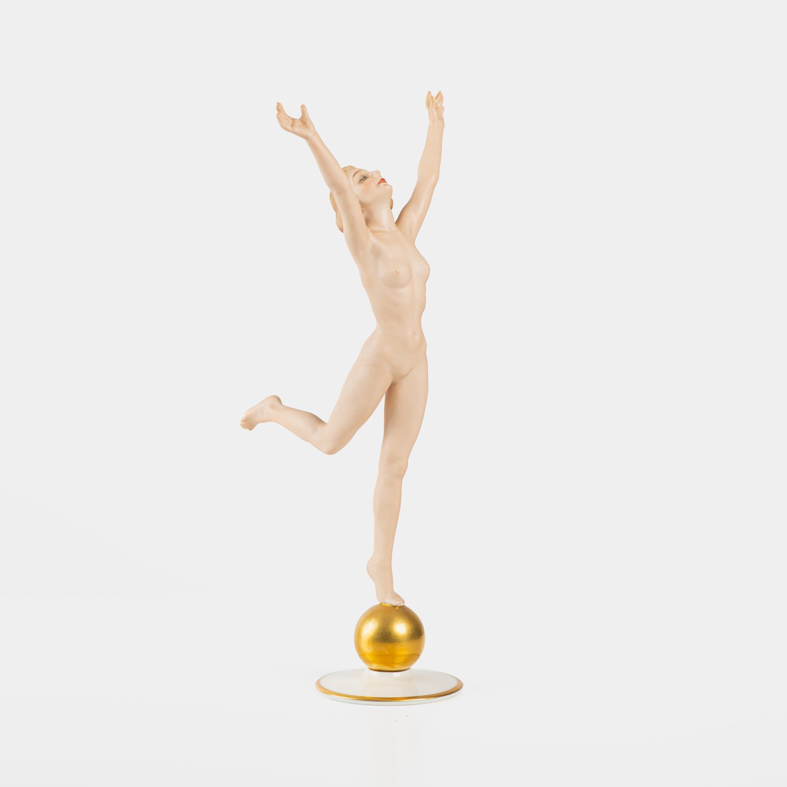 Art Deco porcelain figurine titled “Sonnenkind” (“Sun Child”), modeled by Karl Tutter for Hutschenreuther ca. 1935–1940. Depicts a nude young woman balancing on a golden sphere, arms extended in a dynamic pose. White porcelain with a matte pinkish flesh-tone finish, painted facial features, and satin brown hair. Gilded accents on the sphere and base. Rare signed version with circular base and gold trim.
